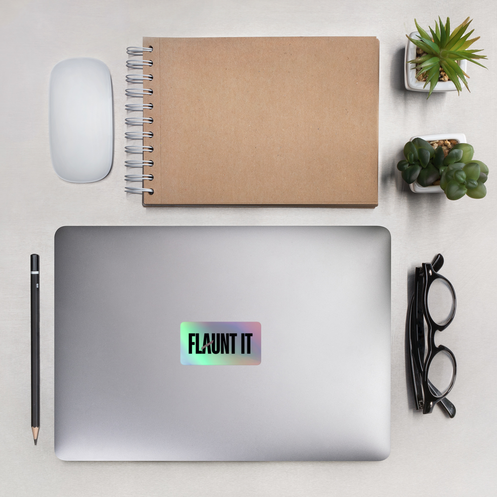 Flaunt It holographic logo sticker on a silver laptop surrounded by a notebook, mouse, pencil, glasses, and plants.