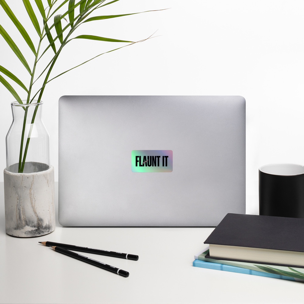 Flaunt It holographic vinyl logo sticker applied to a silver laptop on a desk with a plant and notebooks.