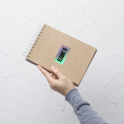 Flaunt It holographic vinyl logo sticker applied to a spiral-bound kraft paper notebook held by a hand
