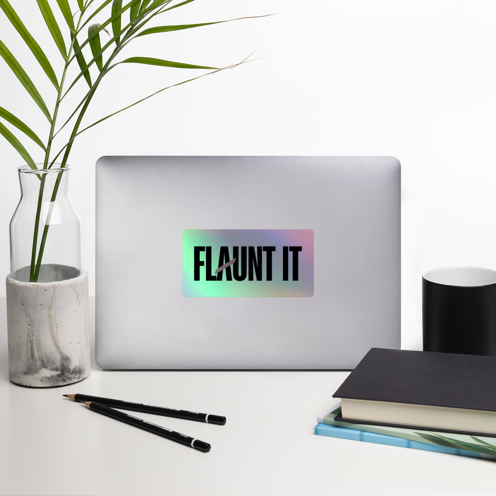 Flaunt It holographic vinyl logo sticker applied to a silver laptop on a desk with a plant and notebook.