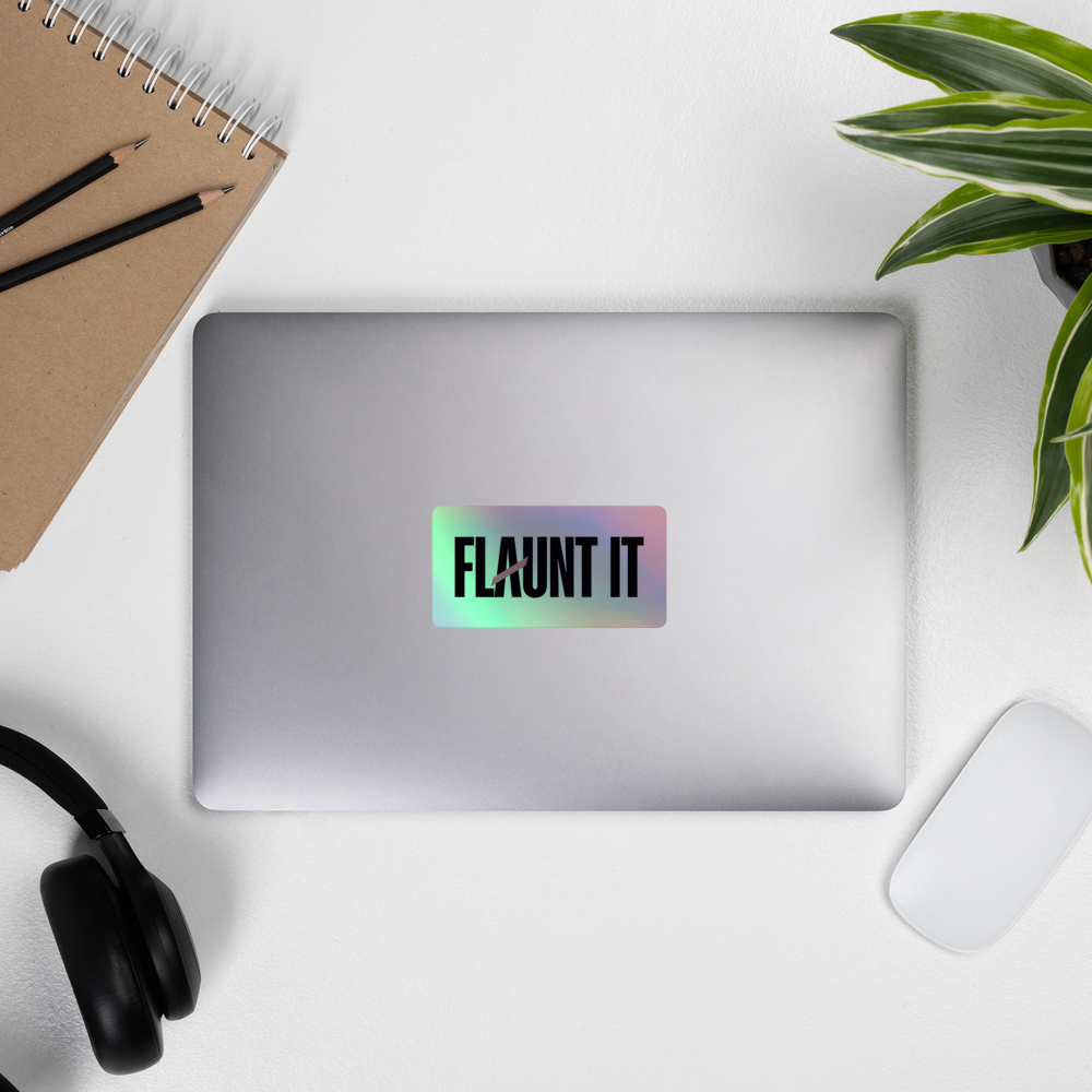 Flaunt It holographic vinyl logo sticker applied to a silver laptop on a desk with a notebook and headphones.