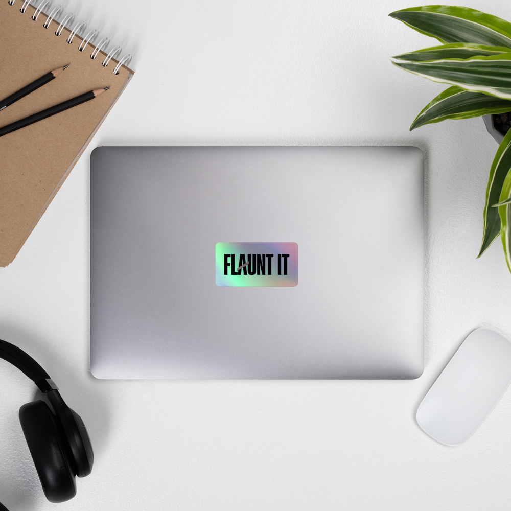 Flaunt It holographic vinyl logo sticker applied to a silver laptop on a desk with a notebook and headphones