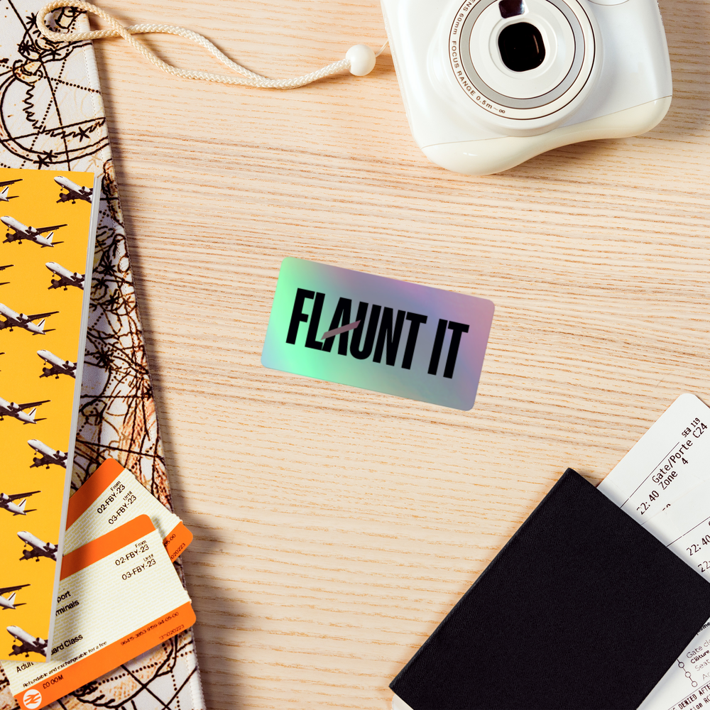 Flaunt It holographic vinyl sticker with 3D embossed logo on a wooden desk with travel accessories.