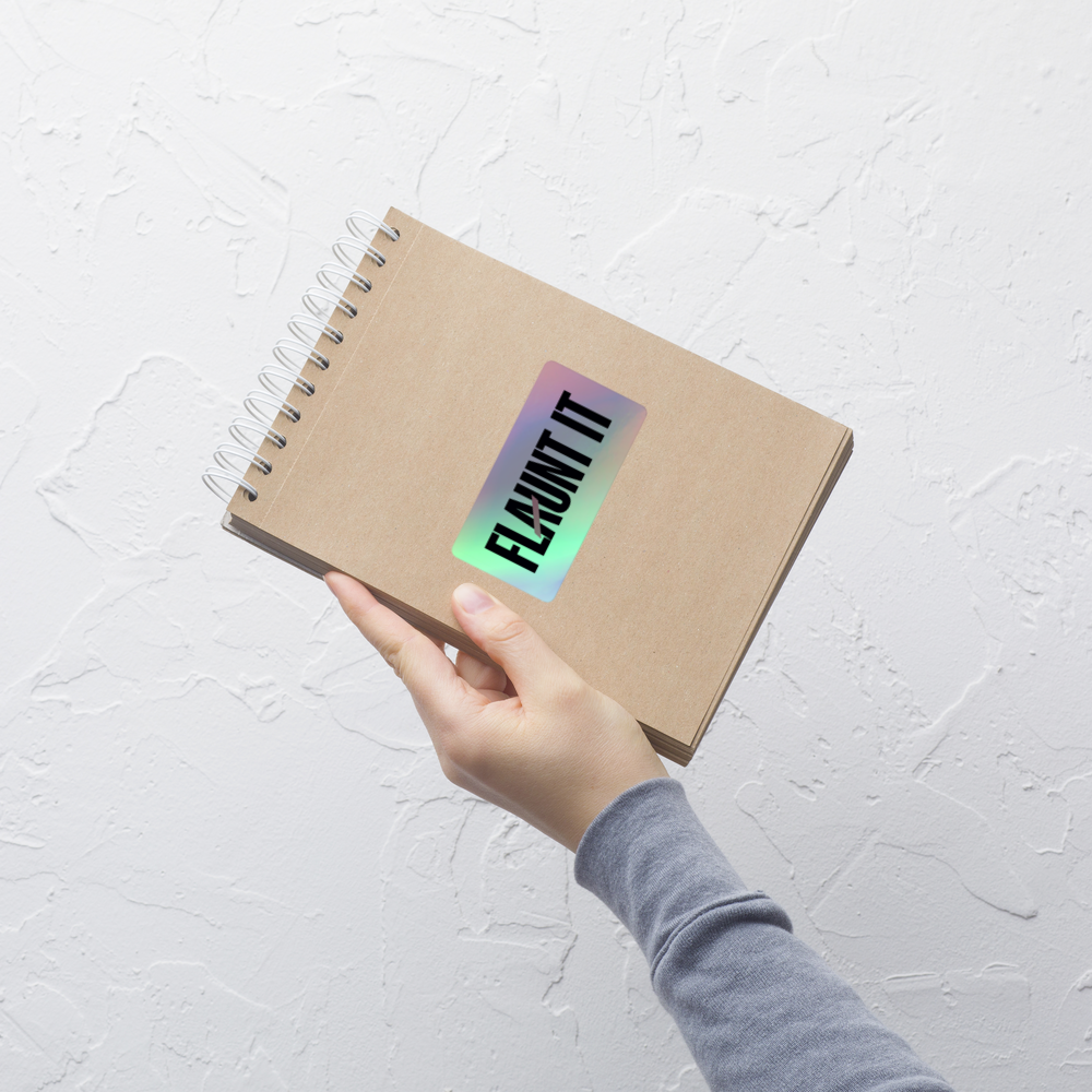 Flaunt It holographic vinyl sticker with 3D logo effect applied to a brown spiral notebook held by a hand