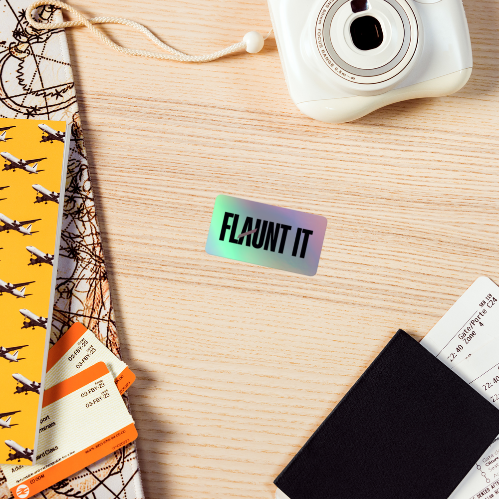 Flaunt It holographic vinyl sticker with a 3D logo effect on a wooden desk with travel accessories