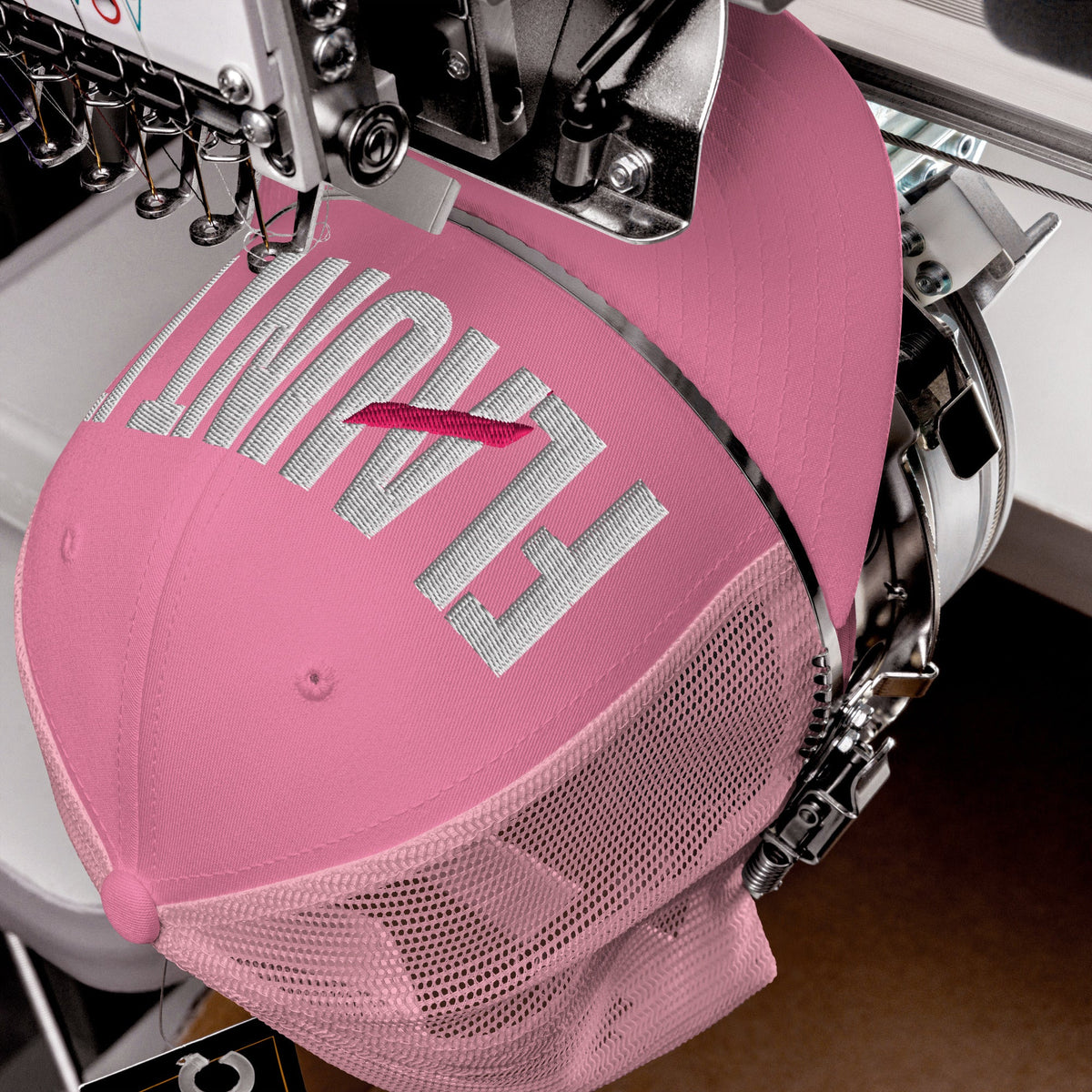 Flaunt It pink mesh-back cap with white embroidered wordmark being stitched by an industrial embroidery machine