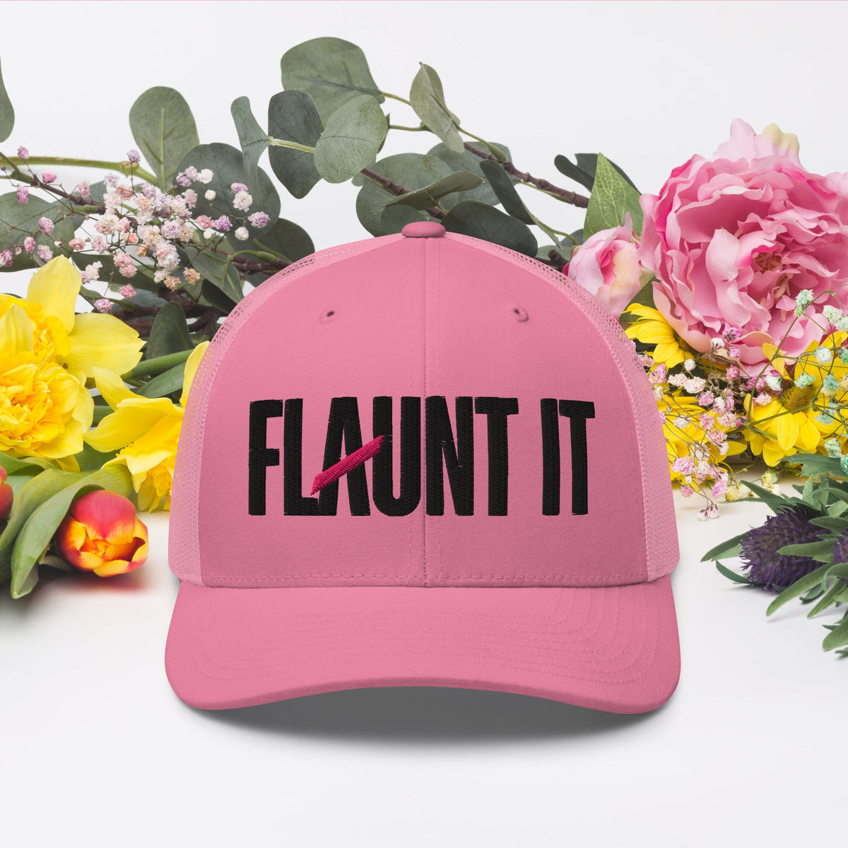 Flaunt It pink six-panel mesh-back cap with black embroidered wordmark logo, styled with colorful spring flowers.