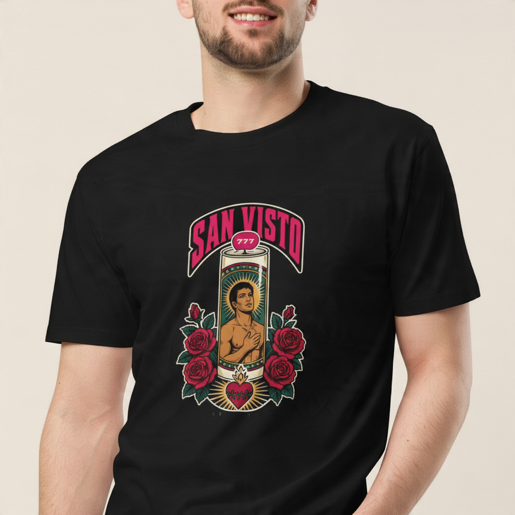 Close-up front view of a smiling male model wearing the black San Visto Premium Unisex T-Shirt with a devotional prayer-candle illustration of a male saint, pink SAN VISTO headline, red roses and a sacred heart, in a warm studio background.