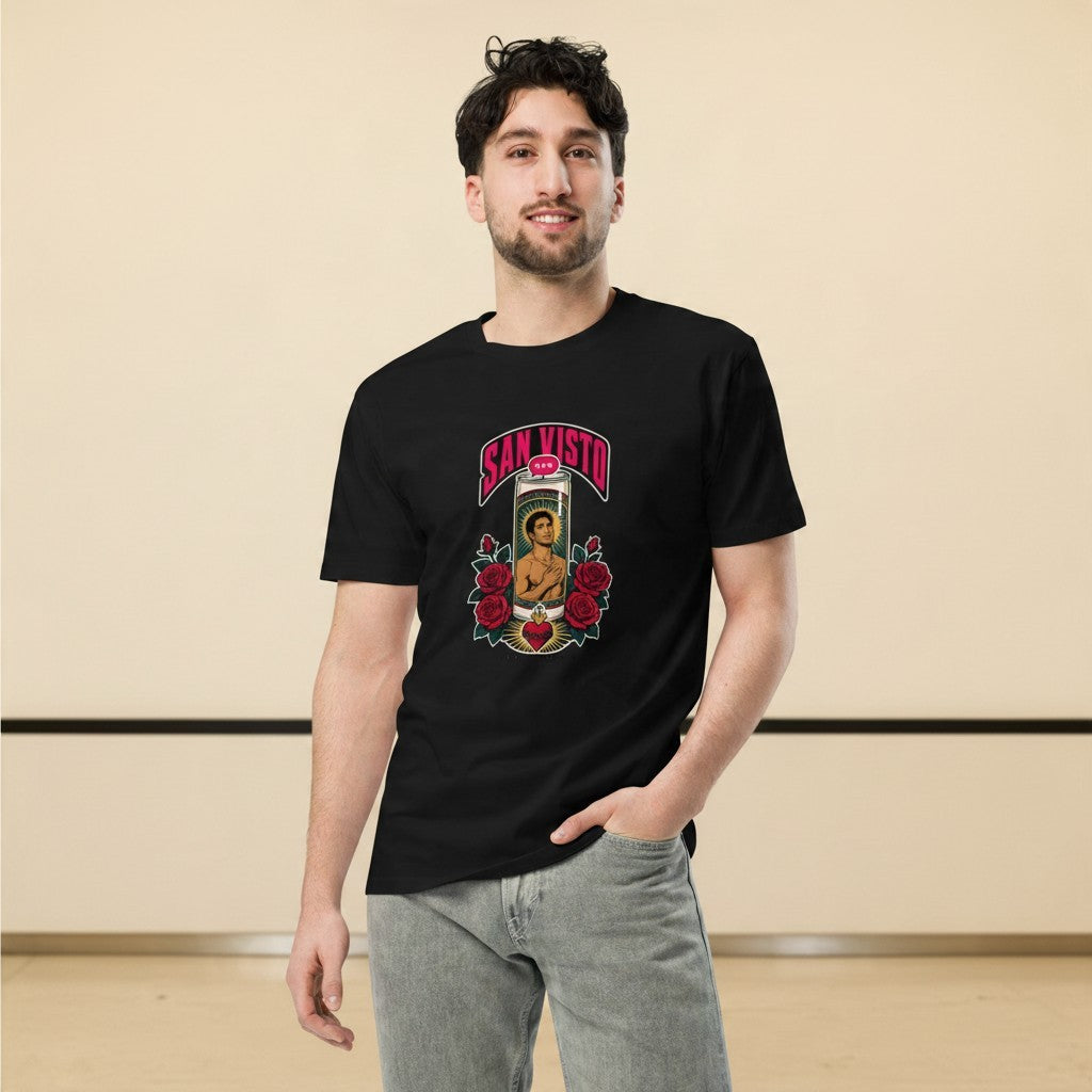 Close-up front view of a smiling male model wearing the black San Visto Premium Unisex T-Shirt with a devotional prayer-candle illustration of a male saint, pink SAN VISTO headline, red roses and a sacred heart, in a warm studio background.