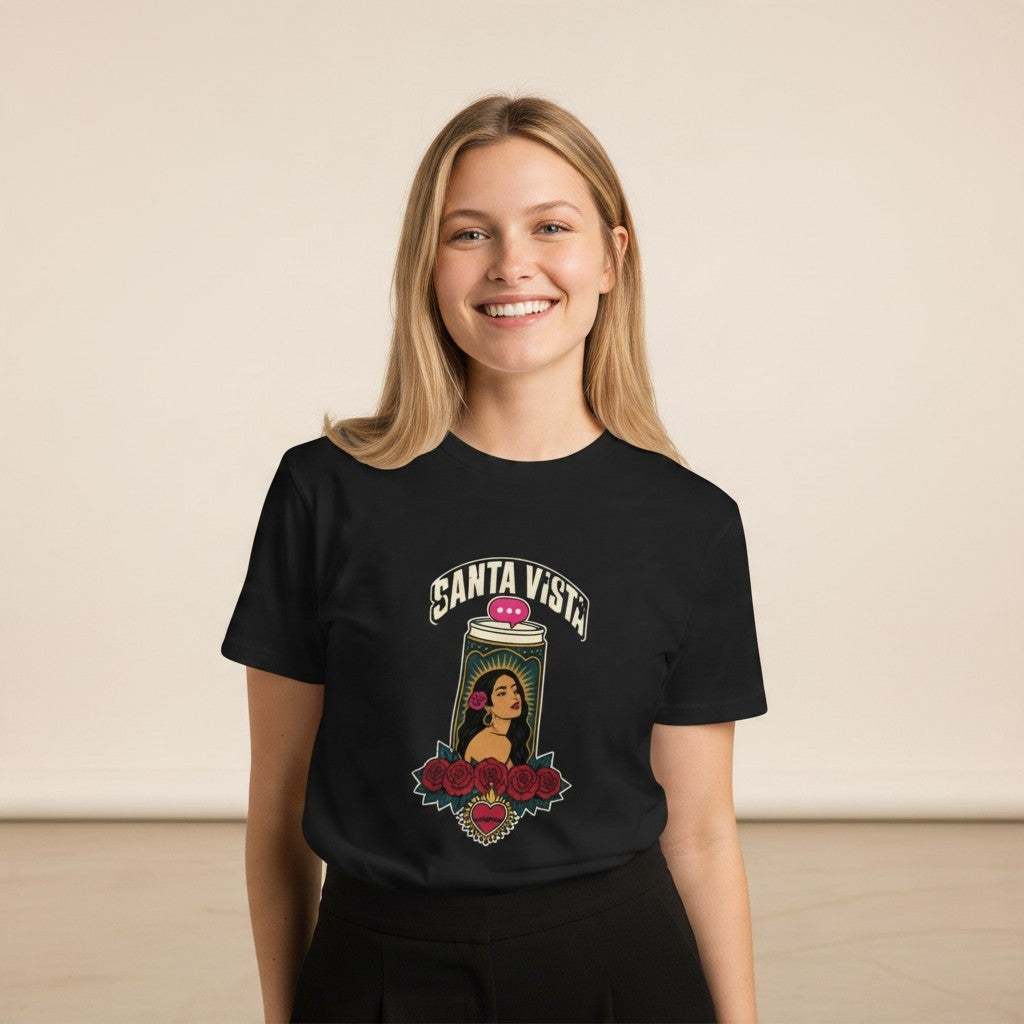 Close-up front view of a smiling female model wearing the black Santa Vista Premium Unisex T-Shirt with a devotional prayer-candle illustration of a female saint, cream SANTA VISTA headline, pink typing bubble, red roses and a sacred heart, in a warm studio background.