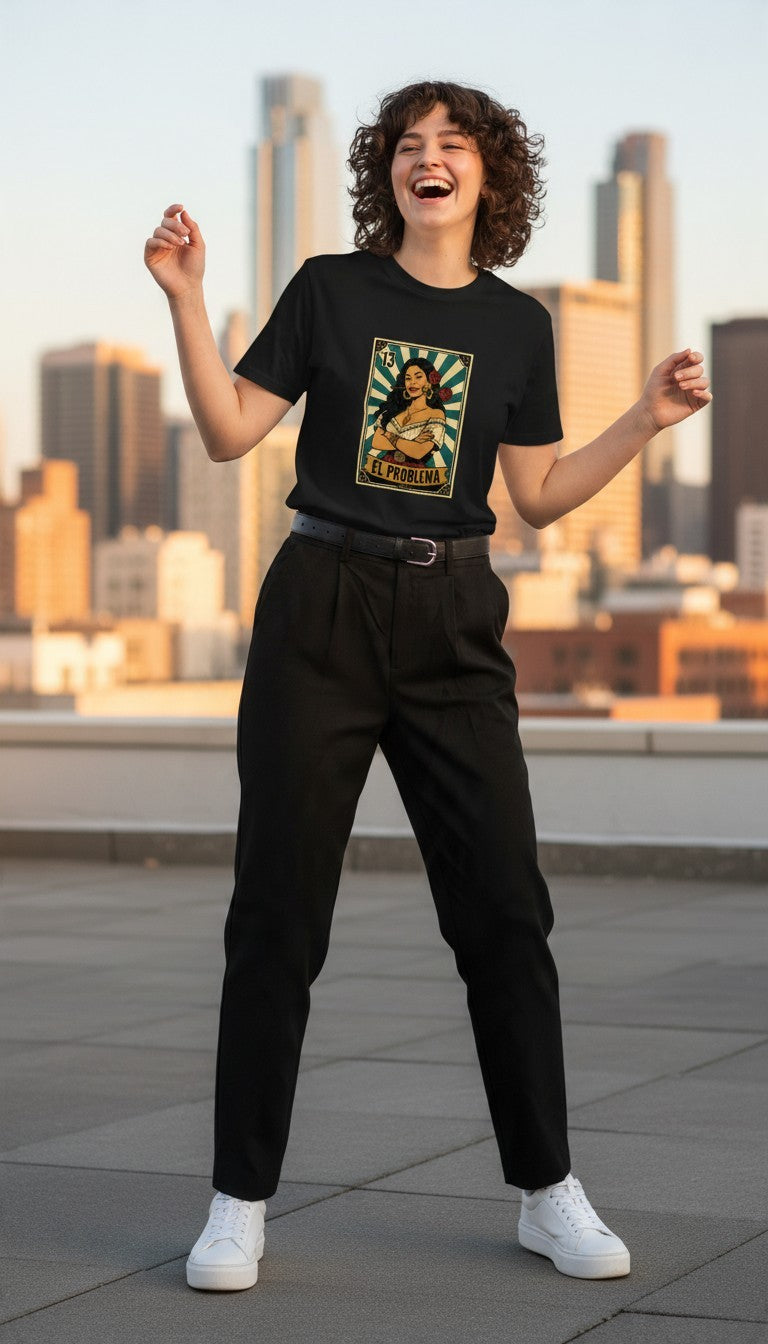 A smiling woman in a black El Problema premium unisex T-shirt with a vintage loteria card design of a woman with roses, EL PROBLEMA text and number 13, laughing on a rooftop at golden hour with a city skyline behind her.