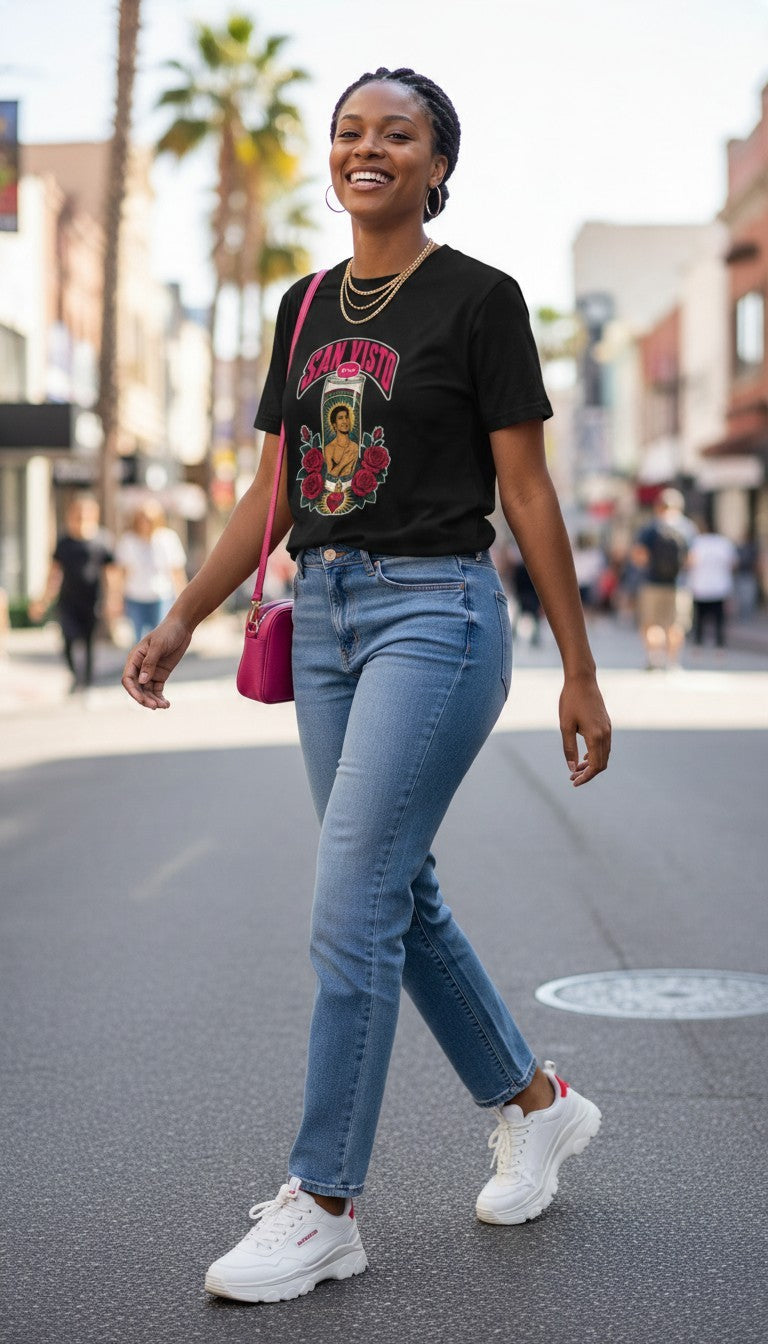 A smiling woman strutting confidently down a sunny city street in three-quarter view, wearing a black San Visto premium unisex T-shirt with a devotional prayer-candle illustration of a male saint, pink SAN VISTO headline, red roses and a sacred heart.