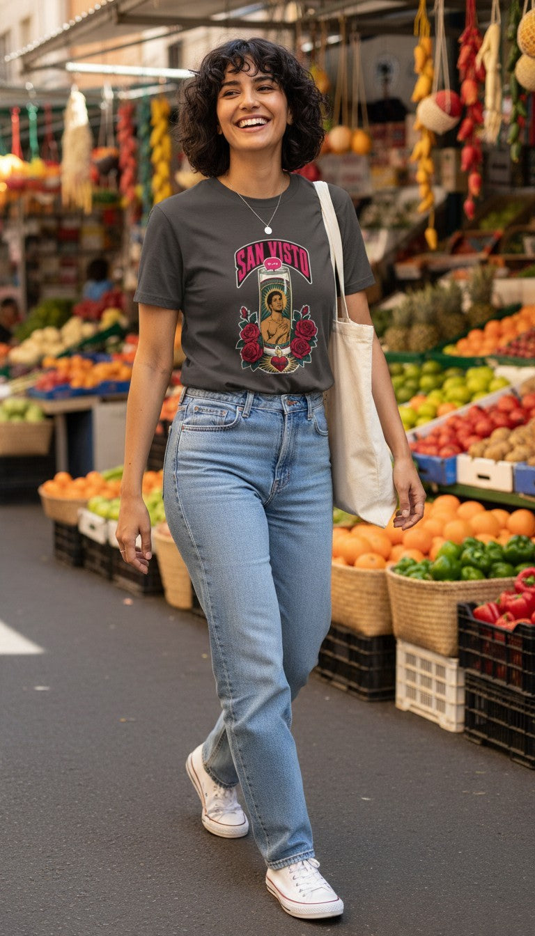 A smiling woman strolling through a colorful outdoor market, wearing a coal San Visto premium unisex T-shirt with a devotional prayer-candle illustration of a male saint, pink SAN VISTO headline, red roses and a sacred heart.