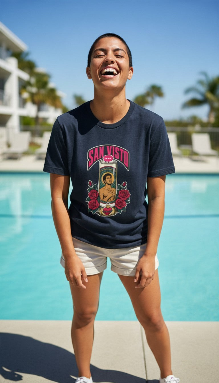 A smiling man laughing by a sunny poolside on a bright summer day, wearing a navy blue San Visto premium unisex T-shirt with a devotional prayer-candle illustration of a male saint, pink SAN VISTO headline, red roses and a sacred heart.