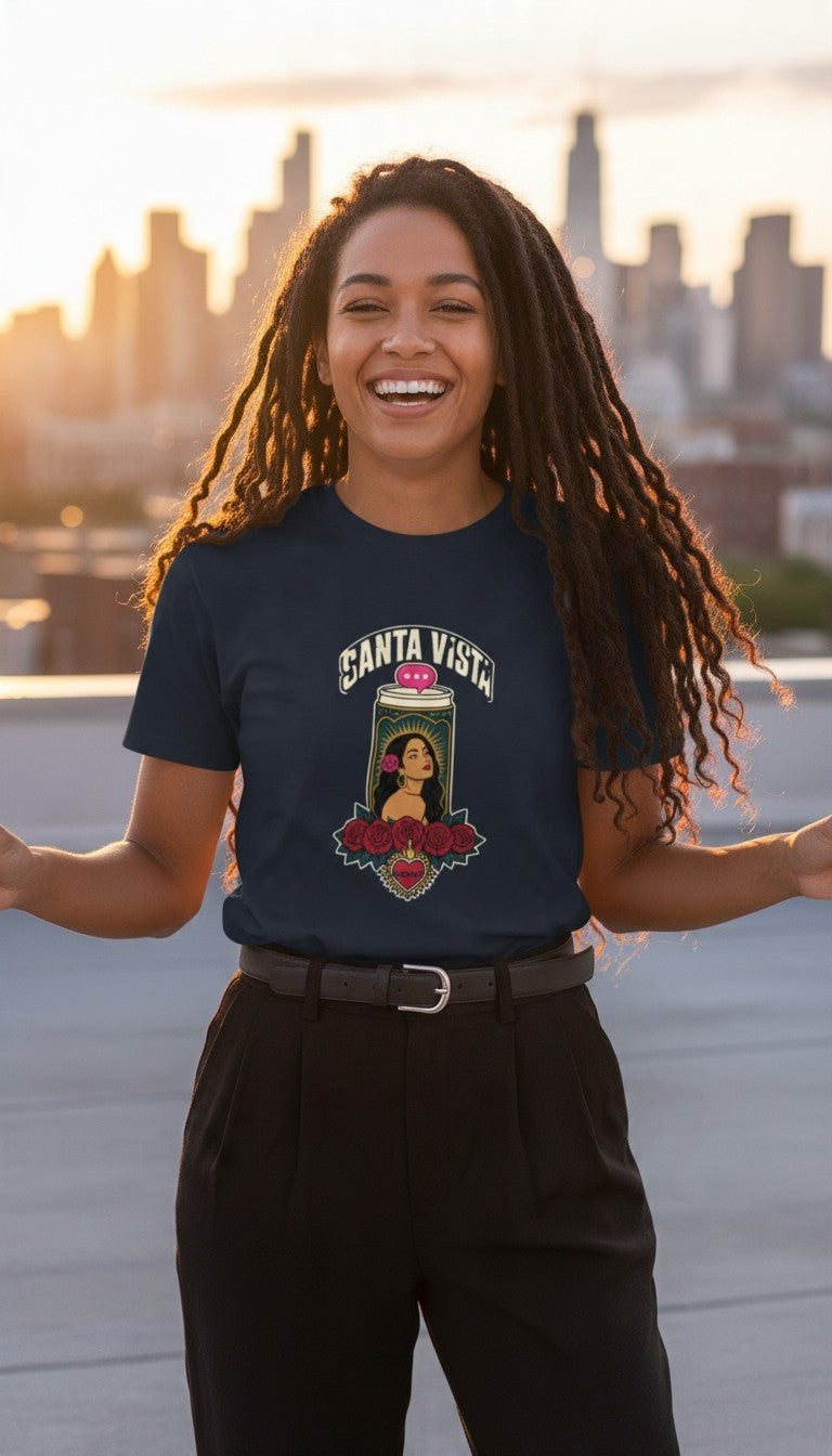 A smiling woman laughing on a rooftop at golden hour with a city skyline behind her, wearing a navy Santa Vista premium unisex T-shirt with a devotional prayer-candle illustration of a female saint, cream SANTA VISTA headline, pink typing bubble, red roses and a sacred heart.