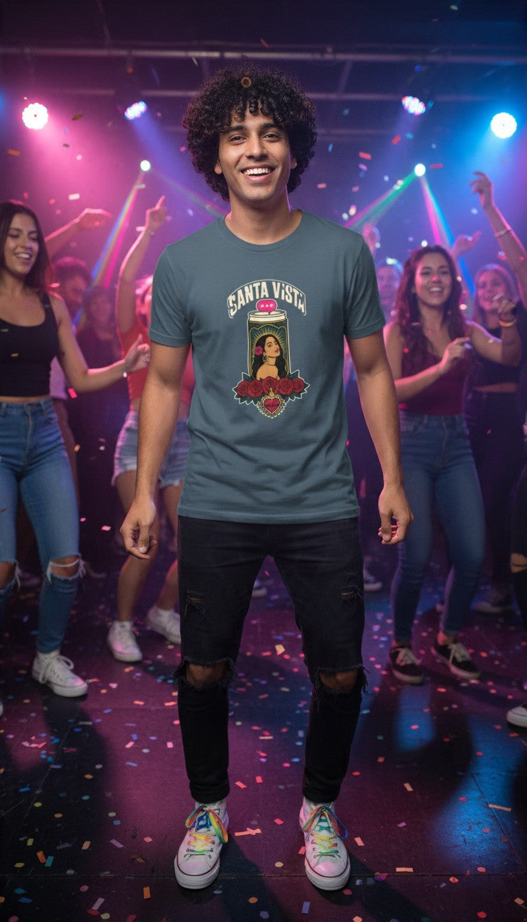 A smiling man dancing joyfully at a fun dance party with friends and colorful lights, wearing a petrol blue Santa Vista premium unisex T-shirt with a devotional prayer-candle illustration of a female saint, cream SANTA VISTA headline, pink typing bubble, red roses and a sacred heart.