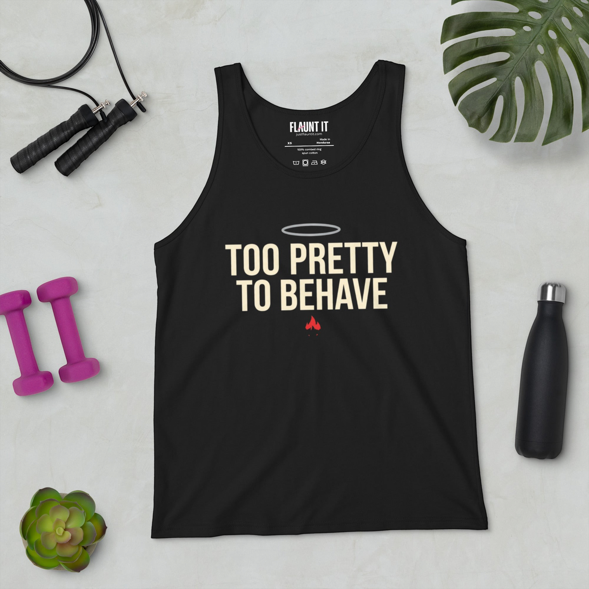 Flaunt It black unisex tank top with Too Pretty to Behave graphic, halo, and flame icon on a flat lay gym background.