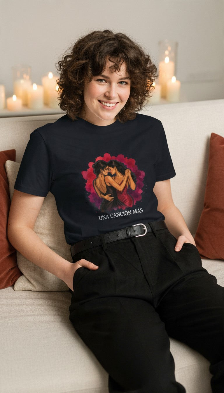 Woman wearing the Una Canción Más Premium Unisex T-Shirt in Navy lounging on a cozy couch surrounded by candles, with the UNA CANCIÓN MÁS watercolor two women dancing design